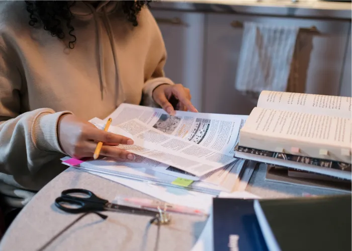 A person reading printed articles and books at a desk, pencil in hand