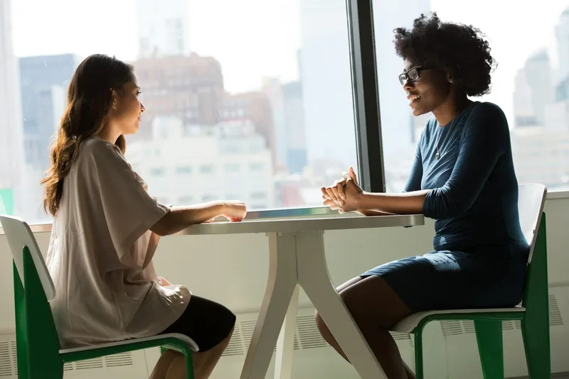 Two people sit at a small table by a window, talking and smiling