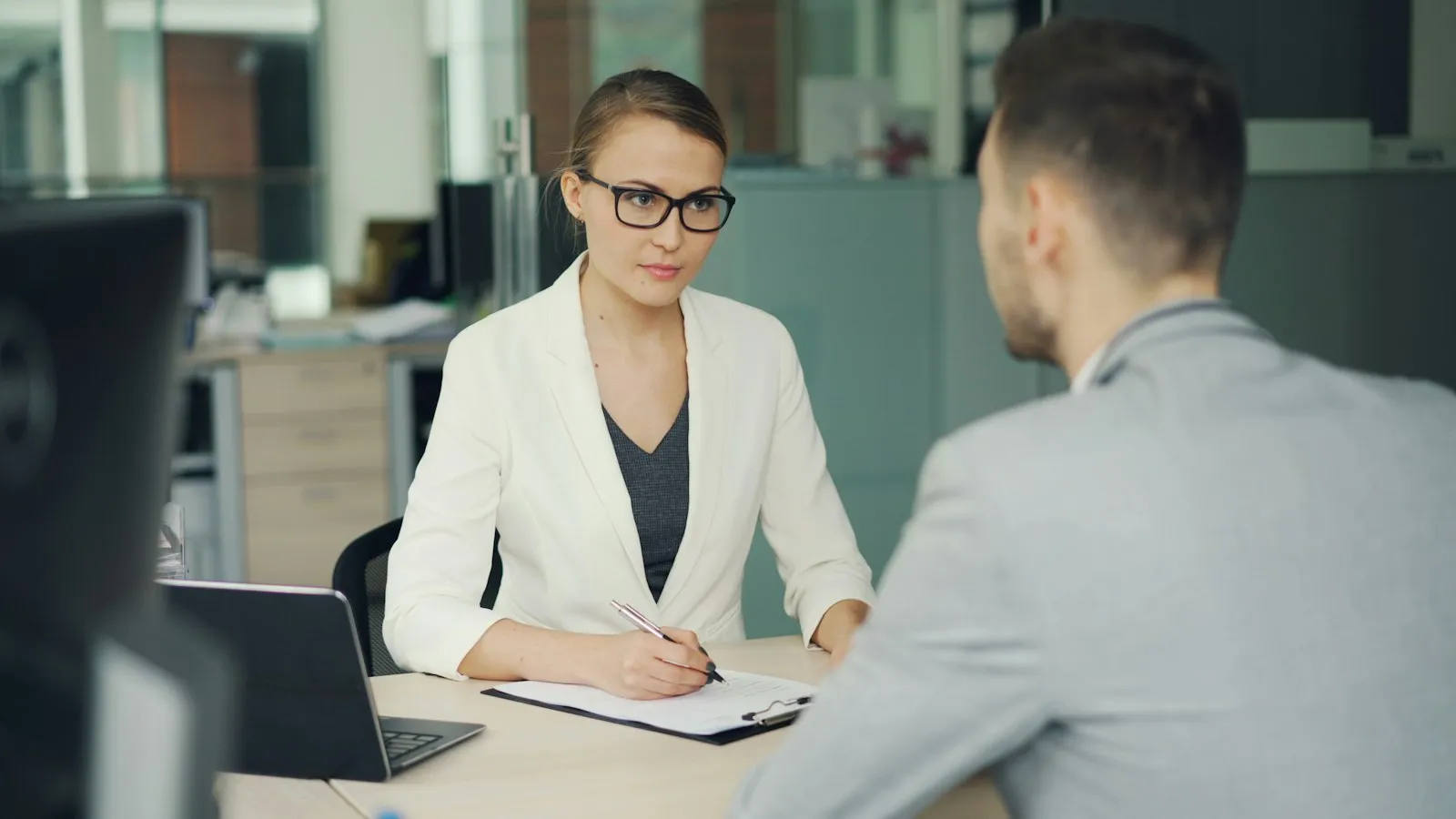 An interviewer writing notes while talking with a candidate at an office desk